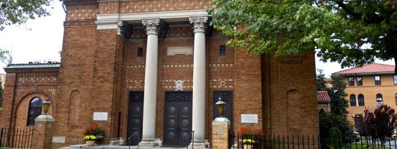 Exterior of a brick building with large stone columns, arched doorway, and decorative brickwork, trees and flowers in front